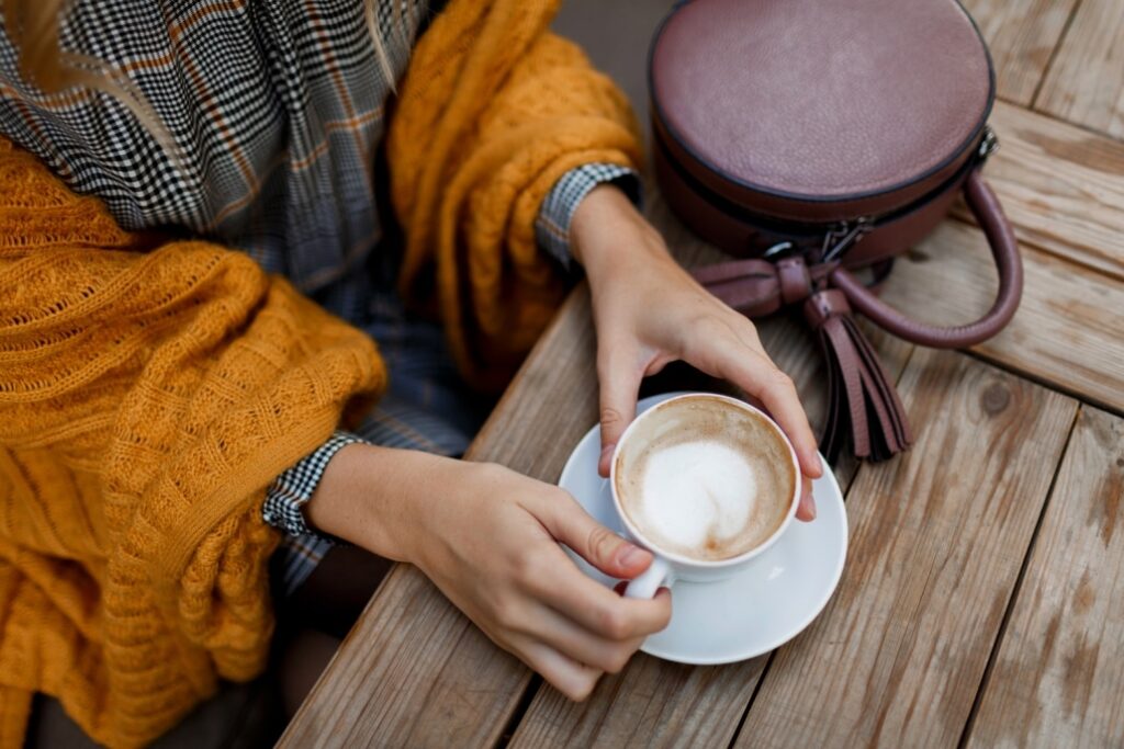 Woman drinking coffee . Stylish bag on table. Wearing grey dress and orange plaid. Enjoying cozy morning in cafe.